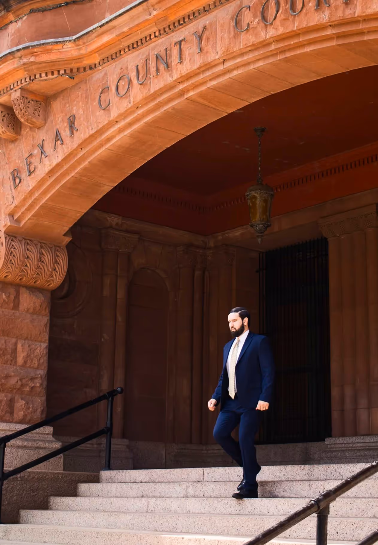 Man in a navy blue suit walking down steps under an arch that reads 'Bexar County Court.'
